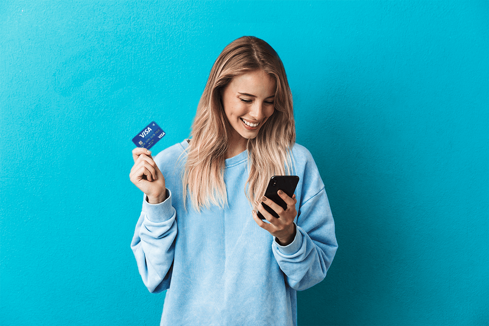 young smiling blond woman dressed in blue jumper holding debit card in right hand and phone in left hand